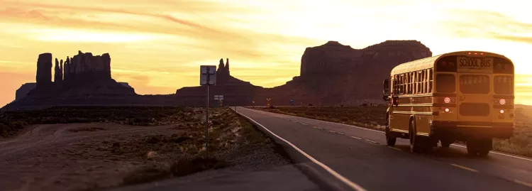 School bus driving into sunsetting over desert mountains.