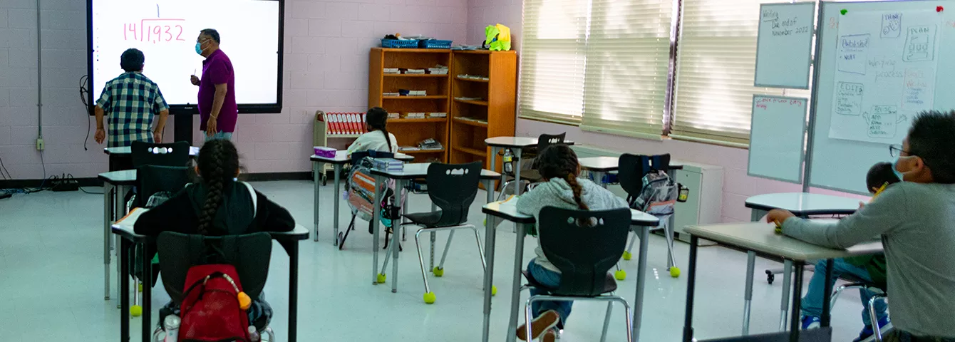 Five students in a classroom watching a male student solving a math problem a the front of the class on a digital board with a male teacher assisting. 