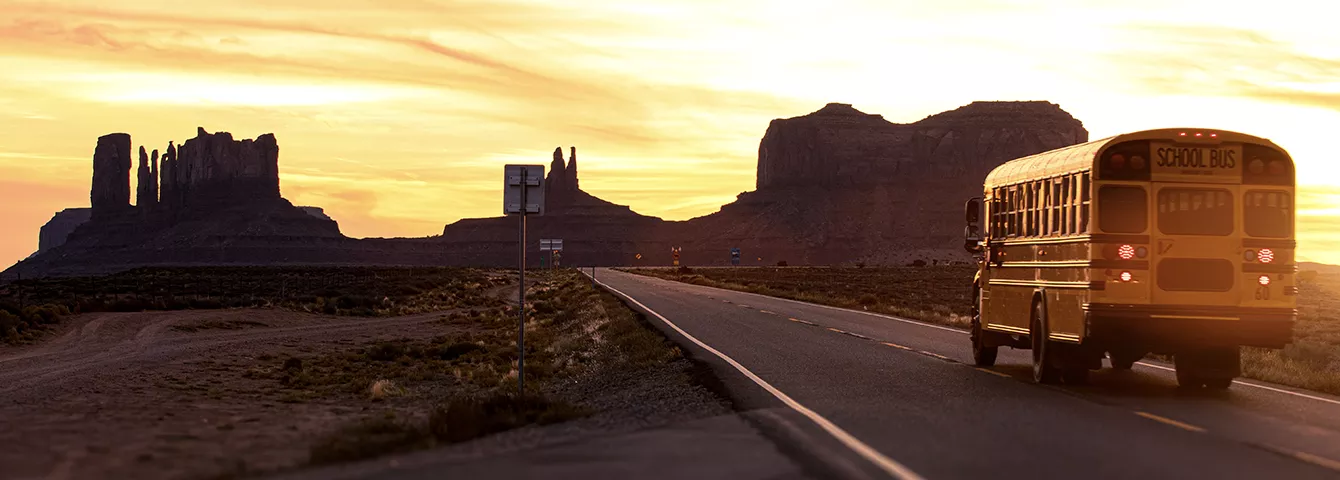 School bus driving into sunsetting over desert mountains.